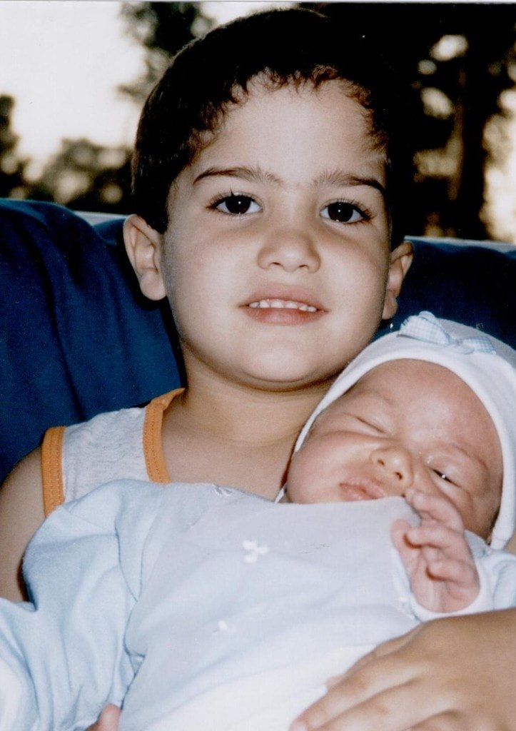 A young Ziv Hagbi carefully cradling his newborn sibling, smiling softly at the camera
