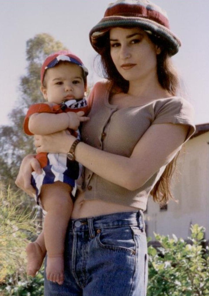 A nostalgic photograph of a woman holding baby Ziv Hagbi outdoors, wearing a striped bucket hat