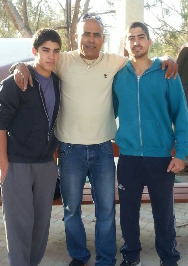 Three generations standing together outdoors, Ziv Hagbi on the right in a blue hoodie, embraced by family