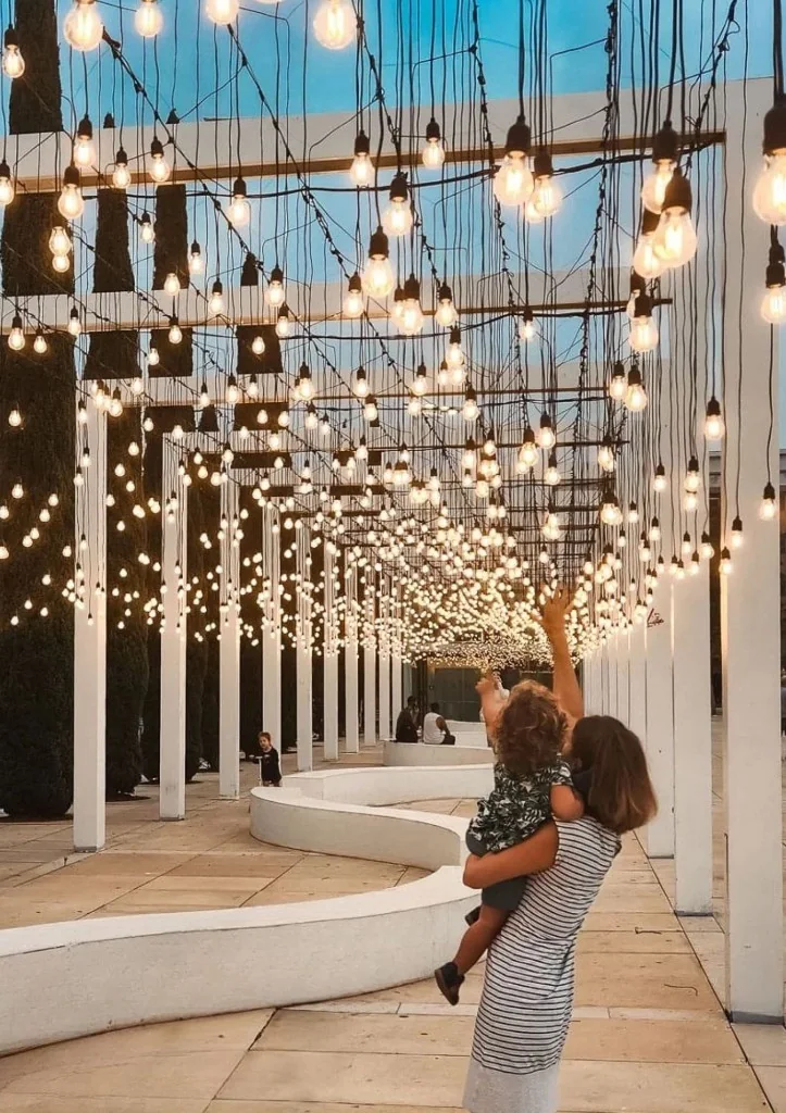 A parent holds a child reaching toward the glowing bulbs at the Light Monument during dusk