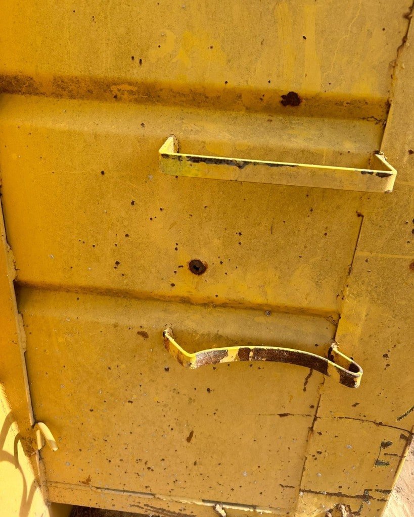 Close-up of a weathered yellow container wall with a small rusted hole between two metal rungs. The lower rung is crushed and bent.