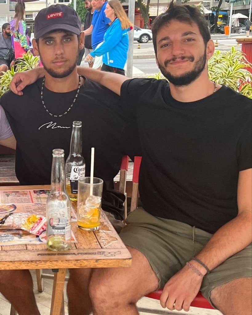 Ron Shemer smiling at an outdoor table beside a man in a Levi's cap, with bottles and a drink on the table