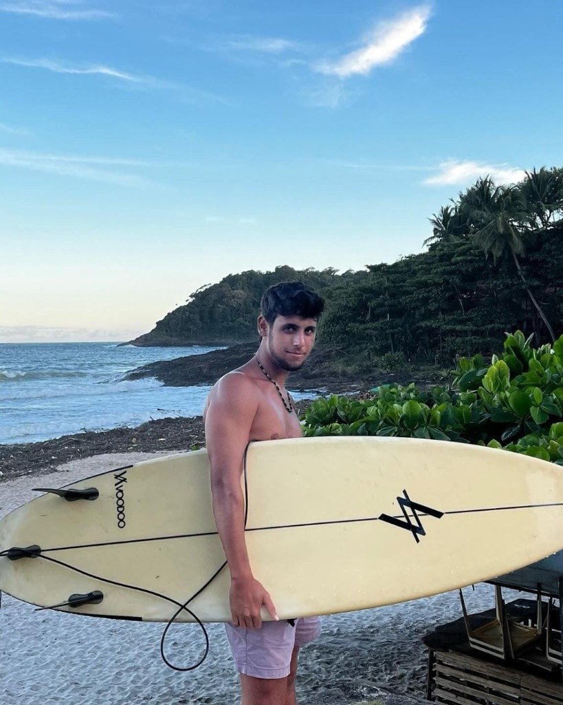 Ron Shemer on a beach holding a pale surfboard, wearing light shorts and a beaded necklace