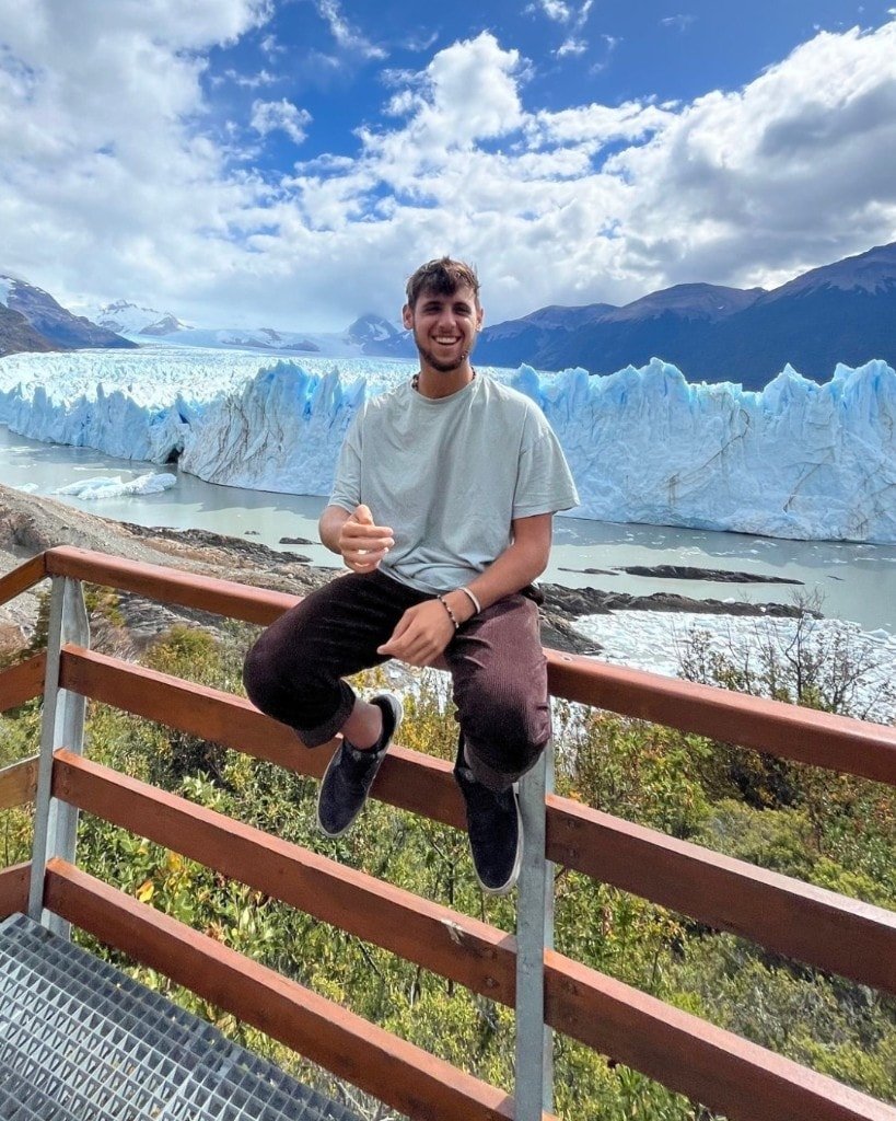 Ron Shemer smiling while sitting on a wooden railing in front of a glacier and mountains