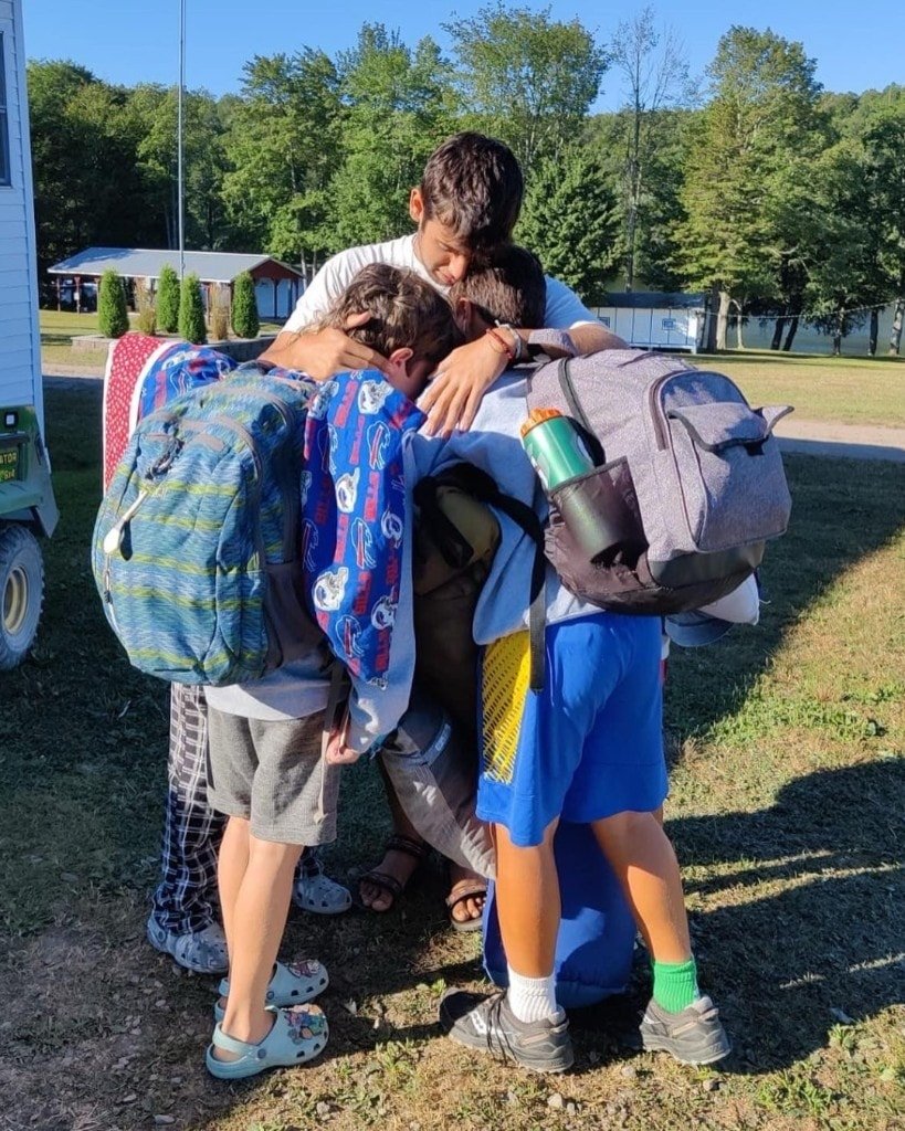 Ron Shemer in a white t-shirt embracing three younger boys wearing backpacks, outdoors on grass