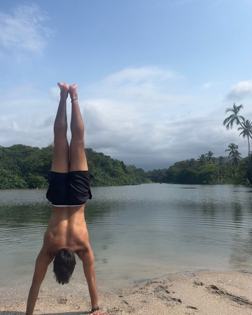 Ron Shemer performing a handstand on a sandy beach beside calm water and trees