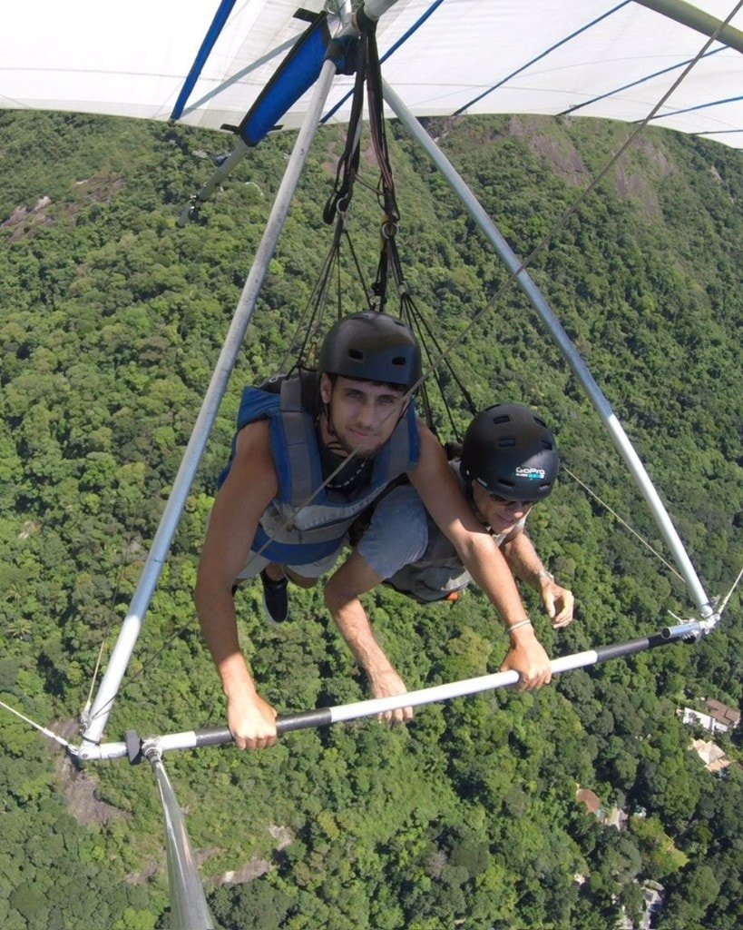 Ron Shemer hang gliding over a forest, wearing a helmet and blue harness beside another flyer